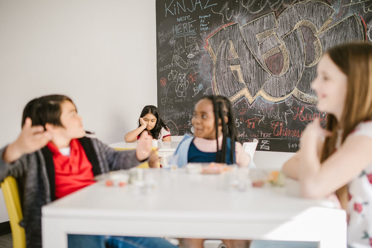 Children gather in a classroom setting with a graffiti-filled chalkboard, one girl sitting apart in focus.