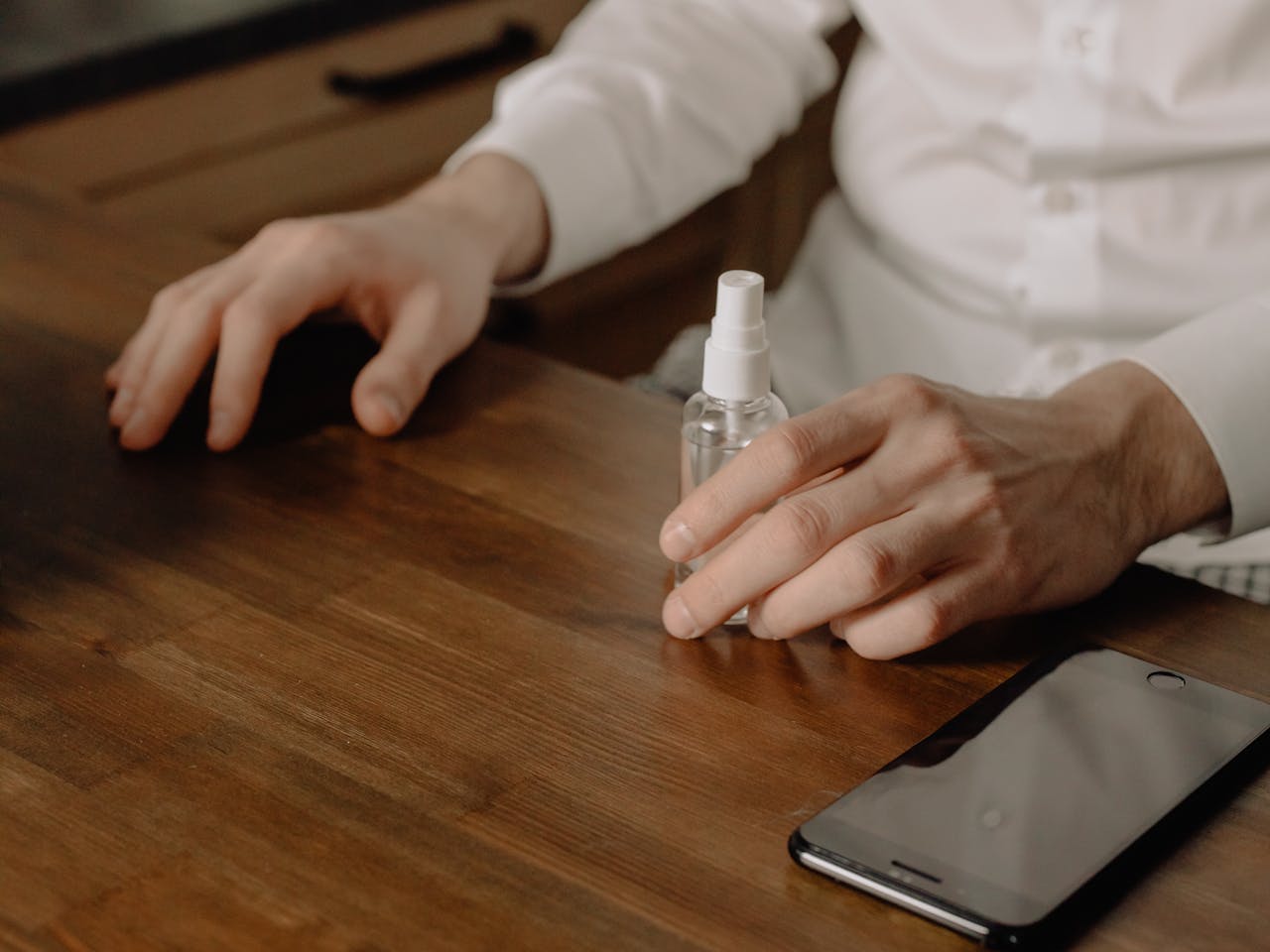 Close-up of hands using sanitizer spray beside a smartphone on a wooden table indoors.